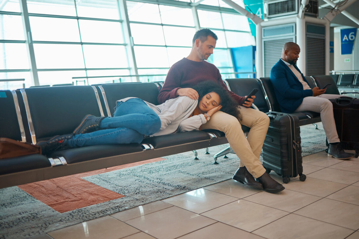 A woman sleeps with her head on a man’s lap while waiting at an airport gate, showing the fatigue and disruption of jet lag after long flights.
