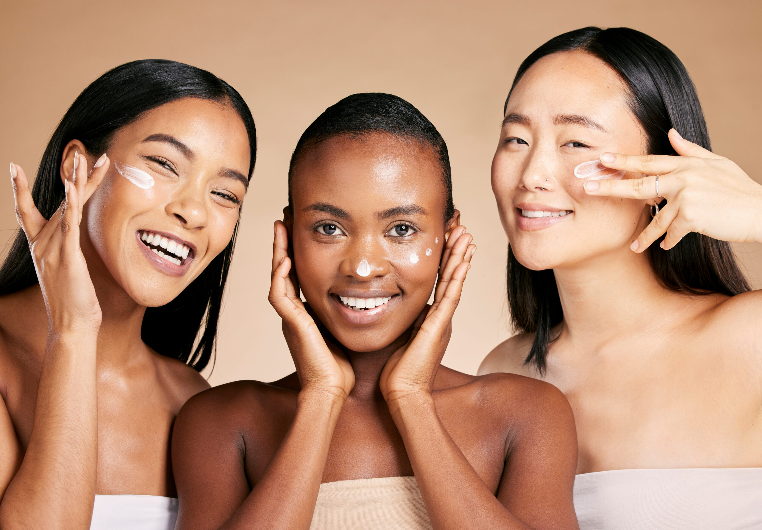 Three women of diverse skin tones smiling while applying facial cream during a skincare routine.