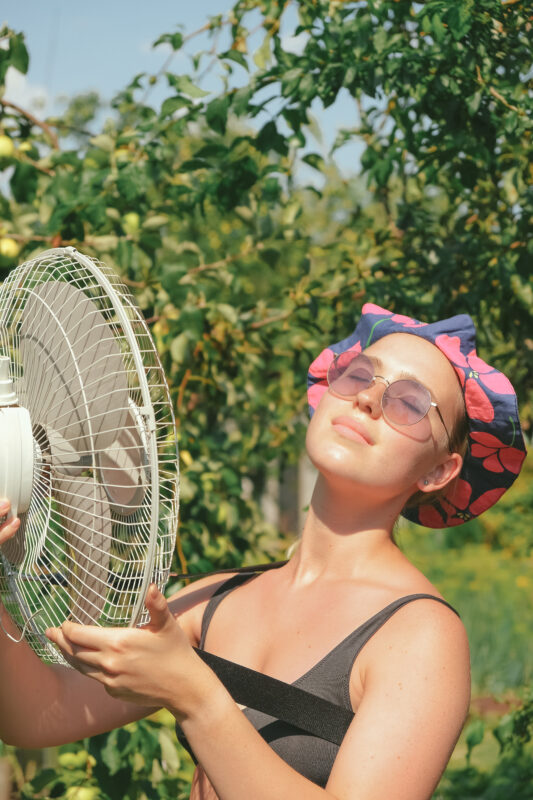 Woman wearing sunglasses using a portable fan outdoors on a hot day, cooling her face in a garden setting during warm summer weather.