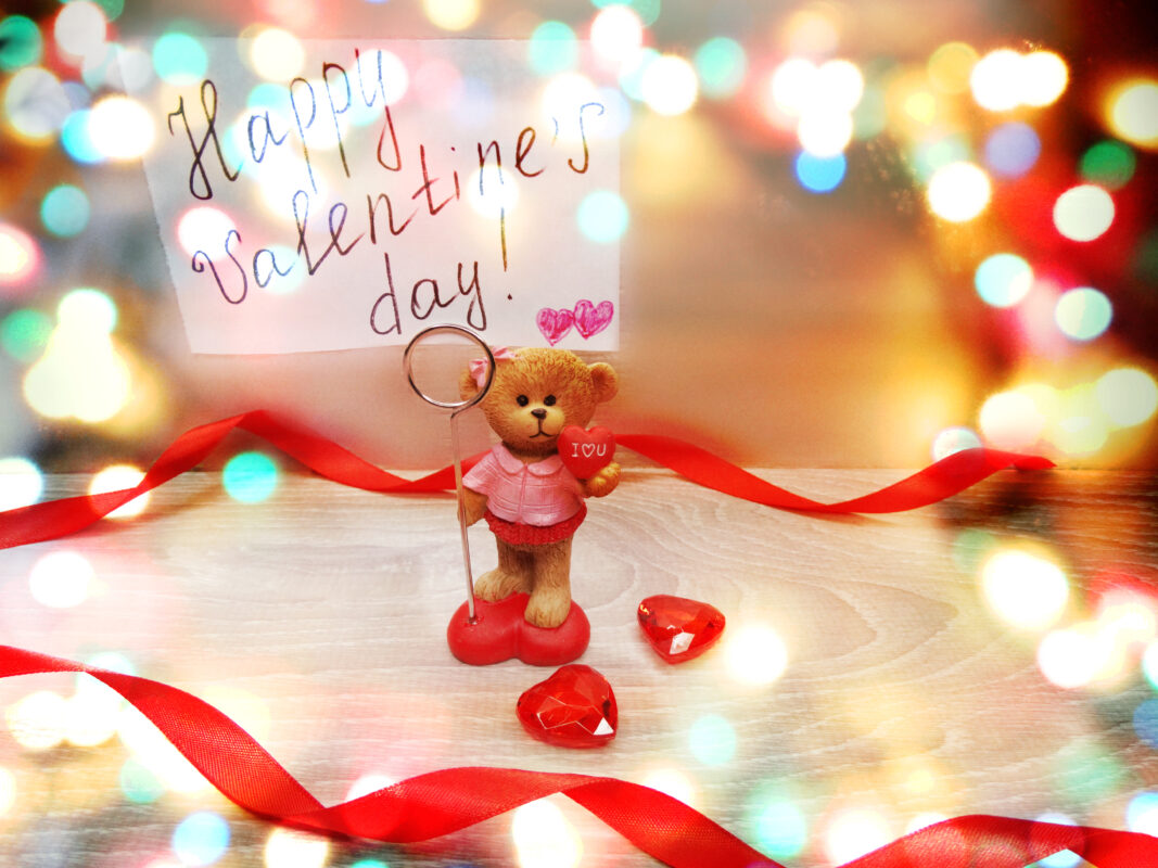Valentine’s Day gift scene with a teddy bear holding a love heart, decorative hearts on a table, and a handwritten Valentine’s card, symbolising thoughtful Valentine’s gifts for her.