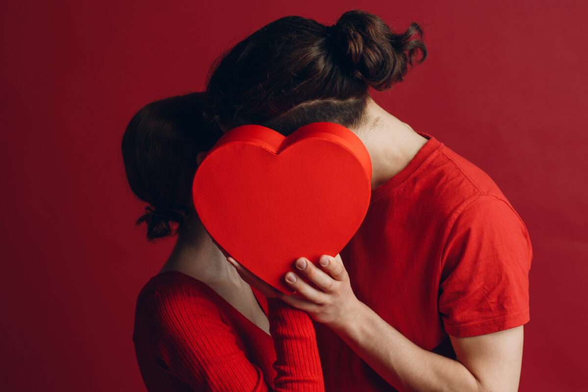 Young couple dressed in red holding a heart-shaped gift box against a red background, representing romantic Valentine’s gifts for her and meaningful gift giving.